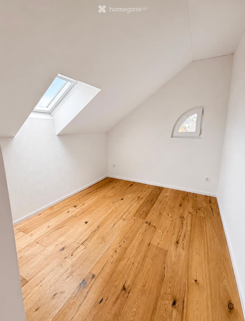 An empty attic room with wooden flooring, white walls, and two windows, one being a skylight.