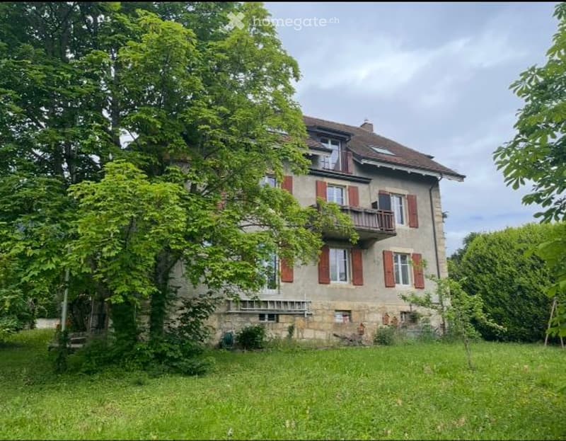 A two-story house with red shutters and a stone facade, surrounded by lush greenery and trees.