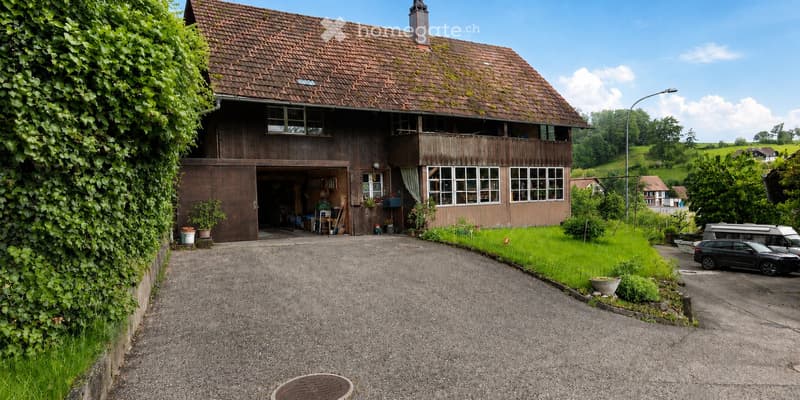 Two-story wooden house with a tiled roof, large windows, and a garage. The house is surrounded by greenery and has a gravel driveway with parked cars.