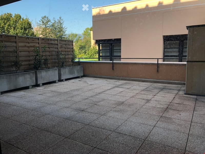 Modern balcony with tiled flooring, low concrete planters with small trees, wooden fence, and a view of greenery and a building.