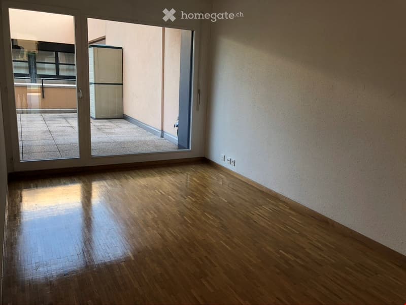 Empty room with wooden flooring, large sliding glass doors leading to a balcony, and a visible refrigerator in the adjacent space.
