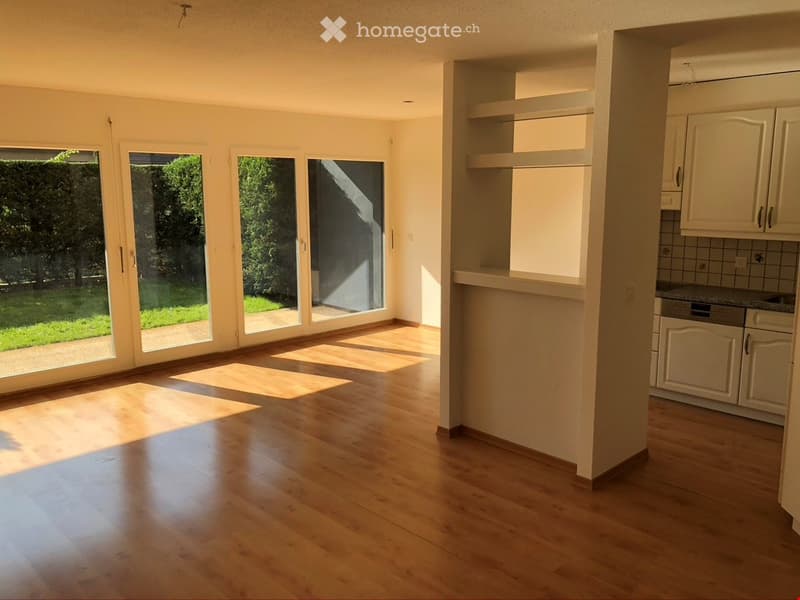 Bright, unfurnished living space with large glass doors leading to a garden, wooden flooring, and an adjacent kitchen area with white cabinets and a countertop.