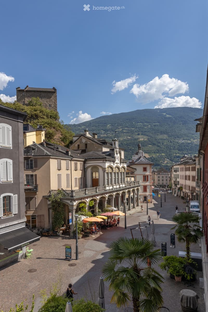 A picturesque town square with cobblestone pavement, outdoor seating under umbrellas, historic buildings, and a scenic mountain backdrop.