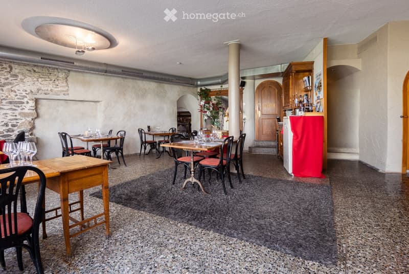 Restaurant interior with wooden tables, chairs, wine glasses, and a refrigerator. The space features a stone wall, carpeted floor, and a wooden door labeled "PRIVE".