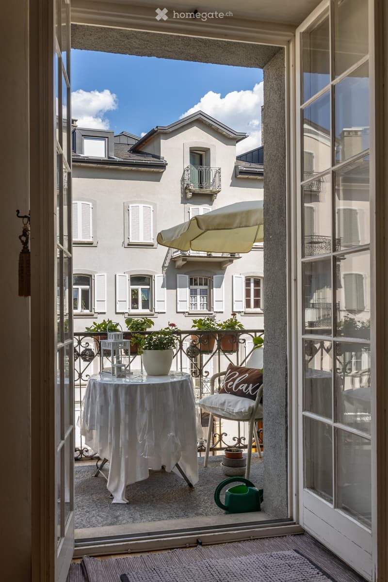 Balcony with a table, chair, potted plants, umbrella, and watering can, overlooking a cityscape with traditional buildings.