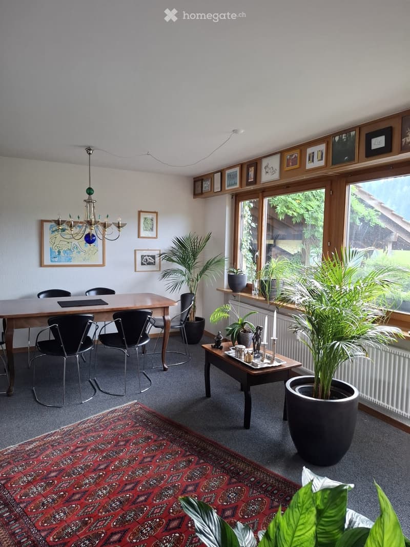 Brightly lit room with large windows, wooden table, black chairs, chandelier, potted plants, and artwork on the walls.