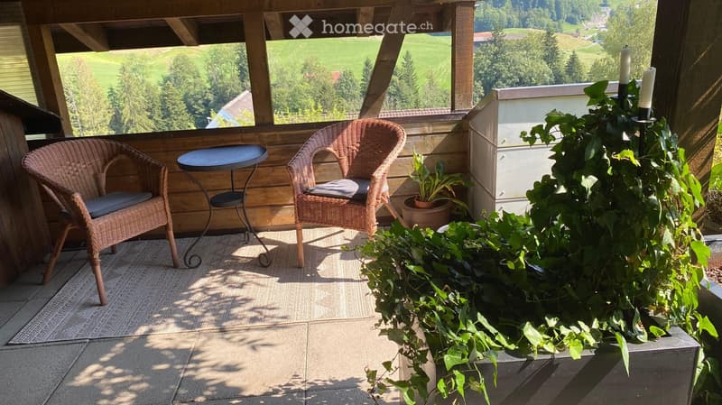 Balcony with wooden beams, wicker chairs, small table, potted plants, and scenic countryside view.