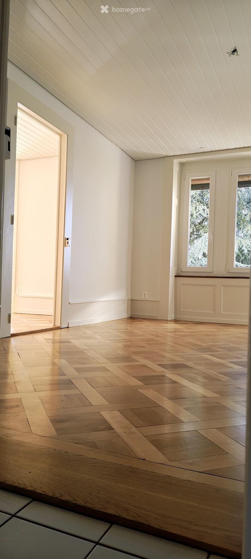 Empty room with wooden herringbone flooring, white walls, and large windows showing outdoor view. No furniture or equipment present.