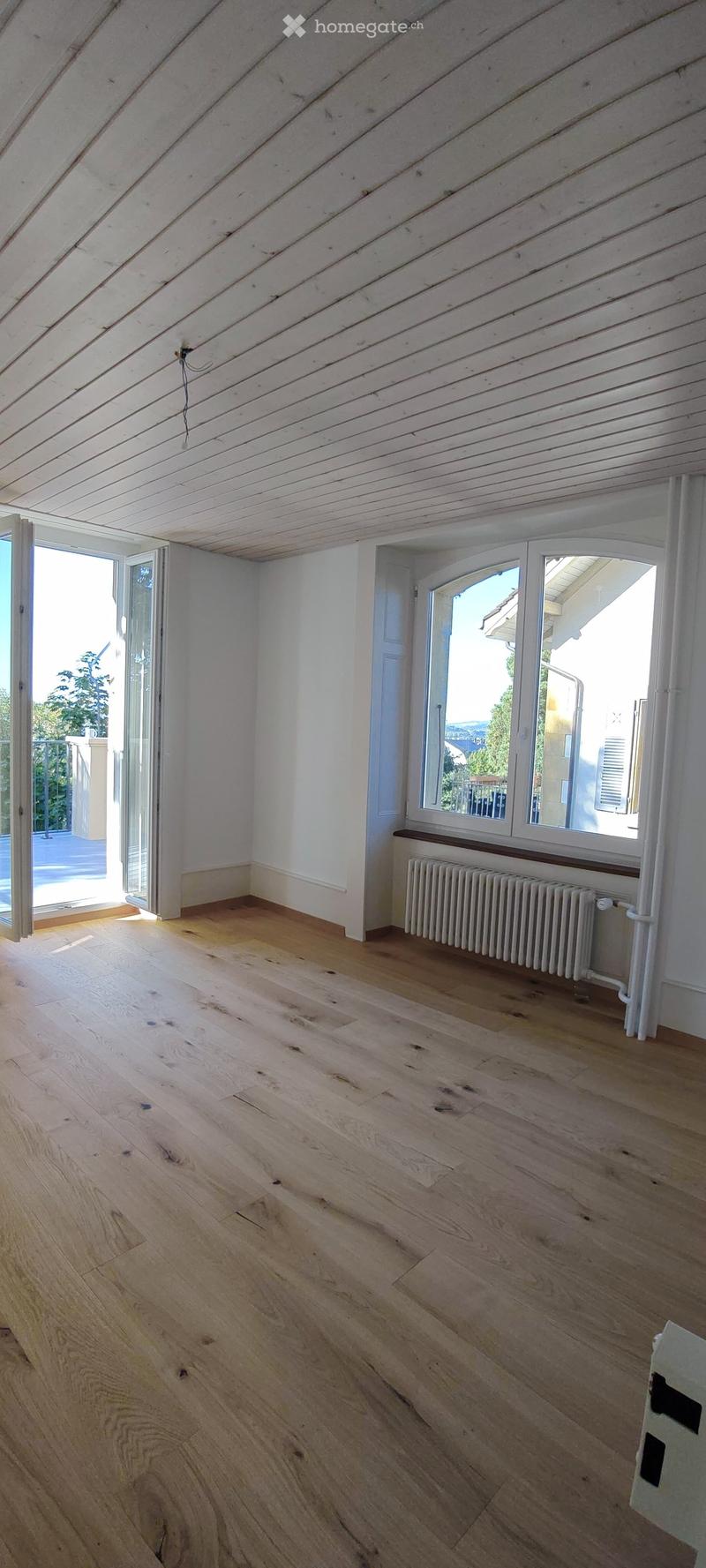 Empty room with wooden flooring, white walls, and large windows with radiators beneath them. The room has exposed wooden beams on the ceiling and an open sliding door leading to a balcony.