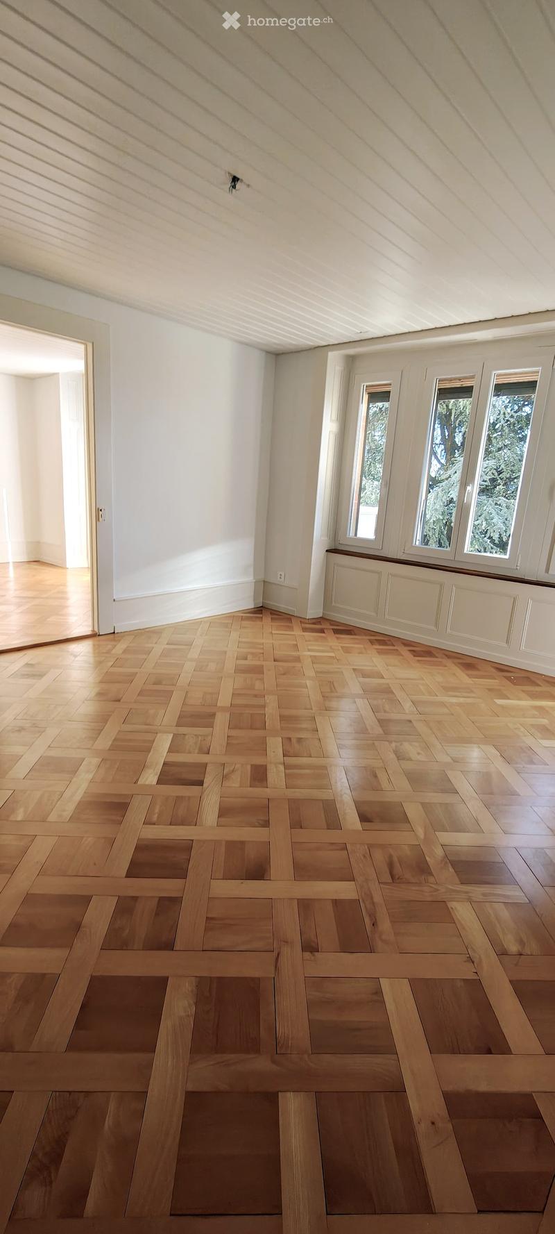 Empty room with wooden herringbone flooring, white walls, and large windows with white frames. The ceiling has exposed wooden beams and a skylight. There is a radiator under the windows and an open doorway leading to another space.