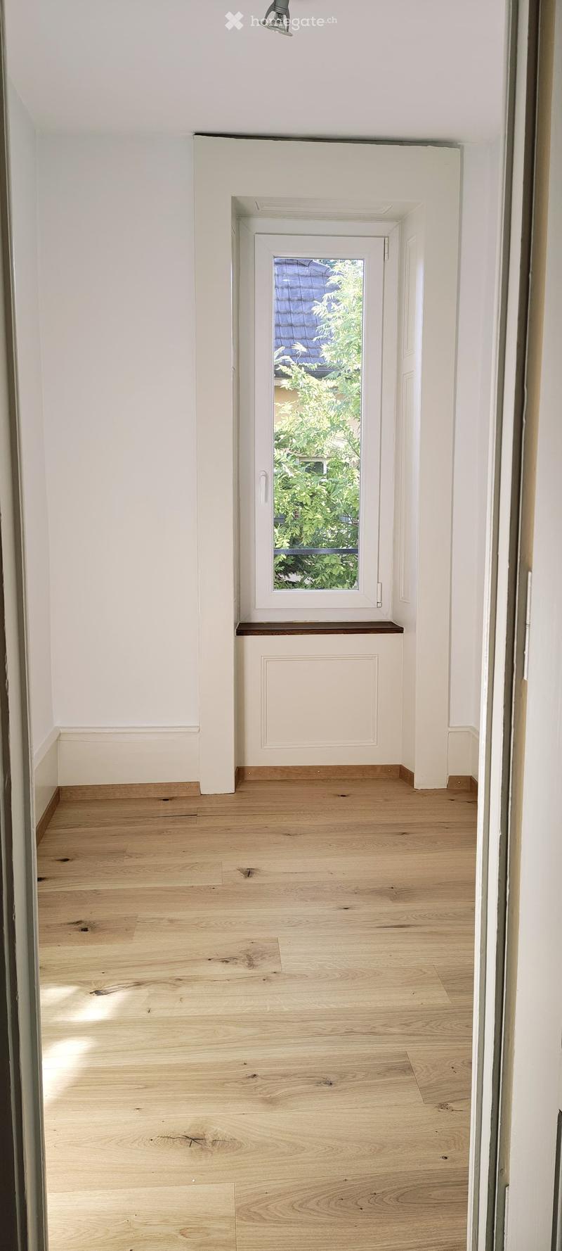 Empty hallway with light wooden flooring, white walls, and a small window showing greenery outside.