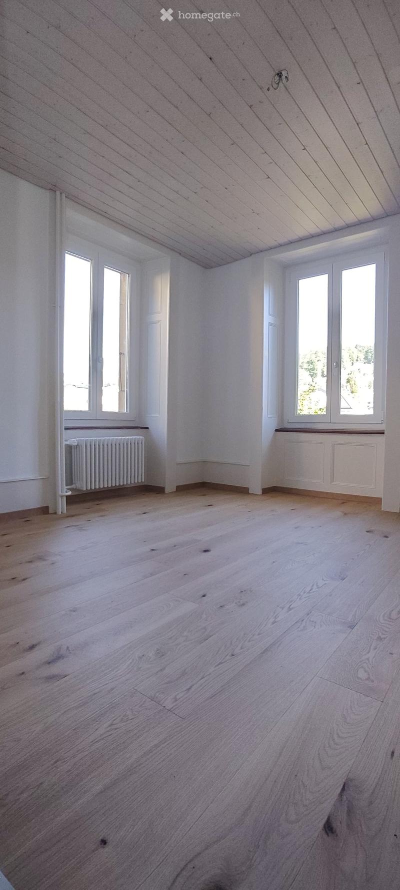 Empty room with wooden flooring, white walls, and large windows allowing natural light. Radiators are visible under the windows.