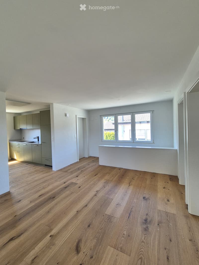 Empty room with light-colored wooden flooring, white walls, and a large window with a view of a neighboring roof. A small kitchen area is visible in the background with cabinets and a sink.
