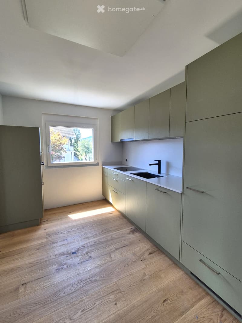 Modern kitchen with light grey cabinets, white countertops, and a double sink. The room features wooden flooring and a window letting in natural light.