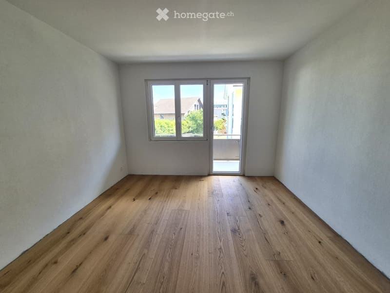 Empty room with light wooden flooring and white walls, featuring a large window and a sliding glass door leading to a balcony.