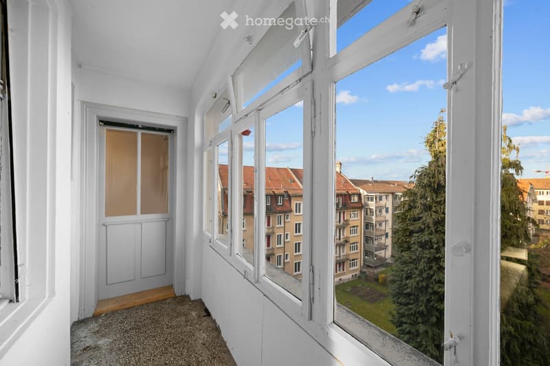 Empty room with large windows offering a view of the city and trees. The room has white walls, a wooden floor, and a door leading to another space.