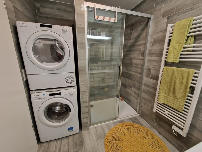 Modern bathroom with a stacked washer and dryer unit, a glass-enclosed shower, a towel rack with green towels, and a yellow crocheted stool.