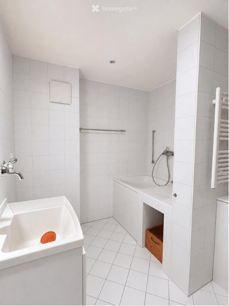 Modern bathroom with white tiled walls and floor, featuring a sink, bathtub, and showerhead.