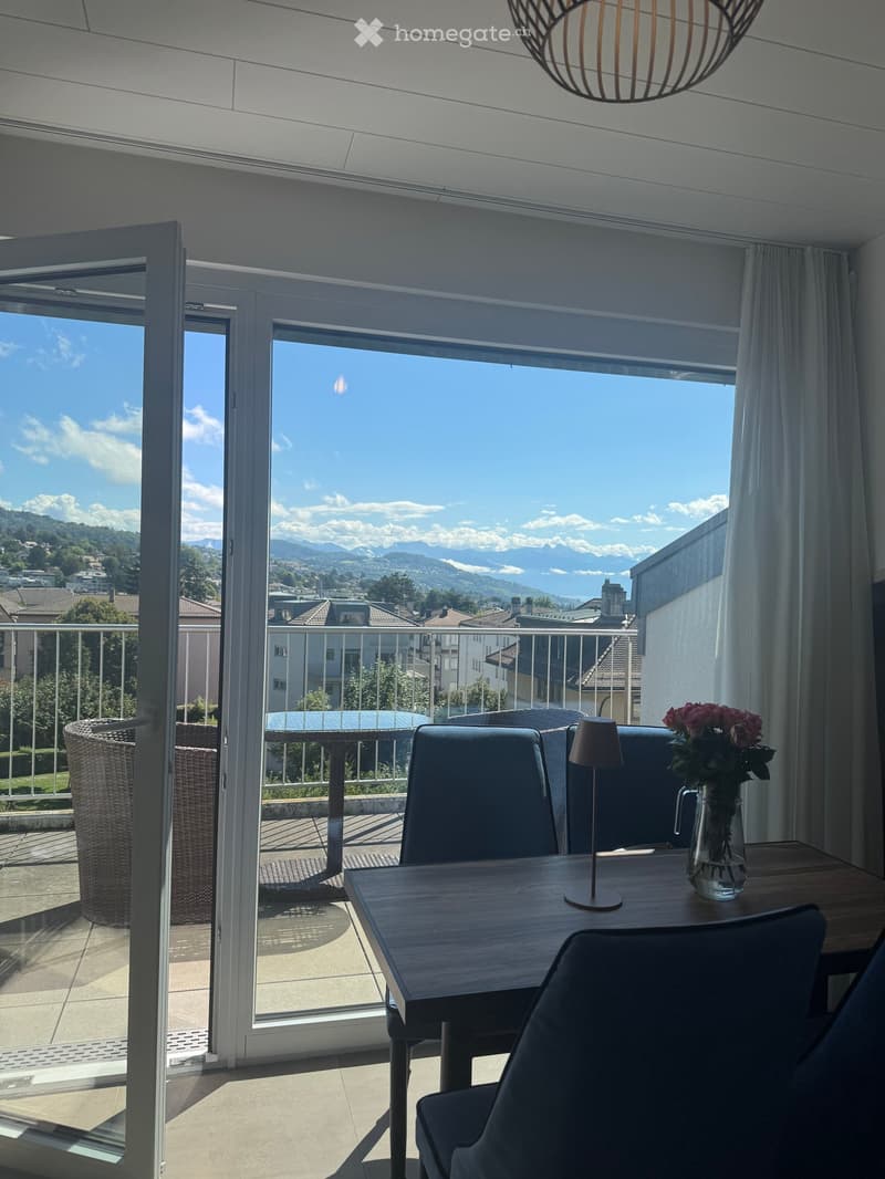Bright room with large windows offering a scenic mountain view, furnished with a wooden table, chairs, a vase with flowers, and a lamp.