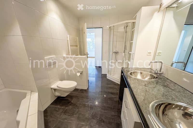 Modern bathroom with white tiles, a bathtub, toilet, double sink with chrome faucets, a large mirror, and a glass-enclosed shower. A radiator is visible on the wall.