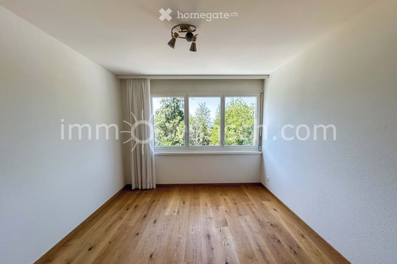 Empty room with wooden flooring, white walls, large window with curtains, and a ceiling light fixture.