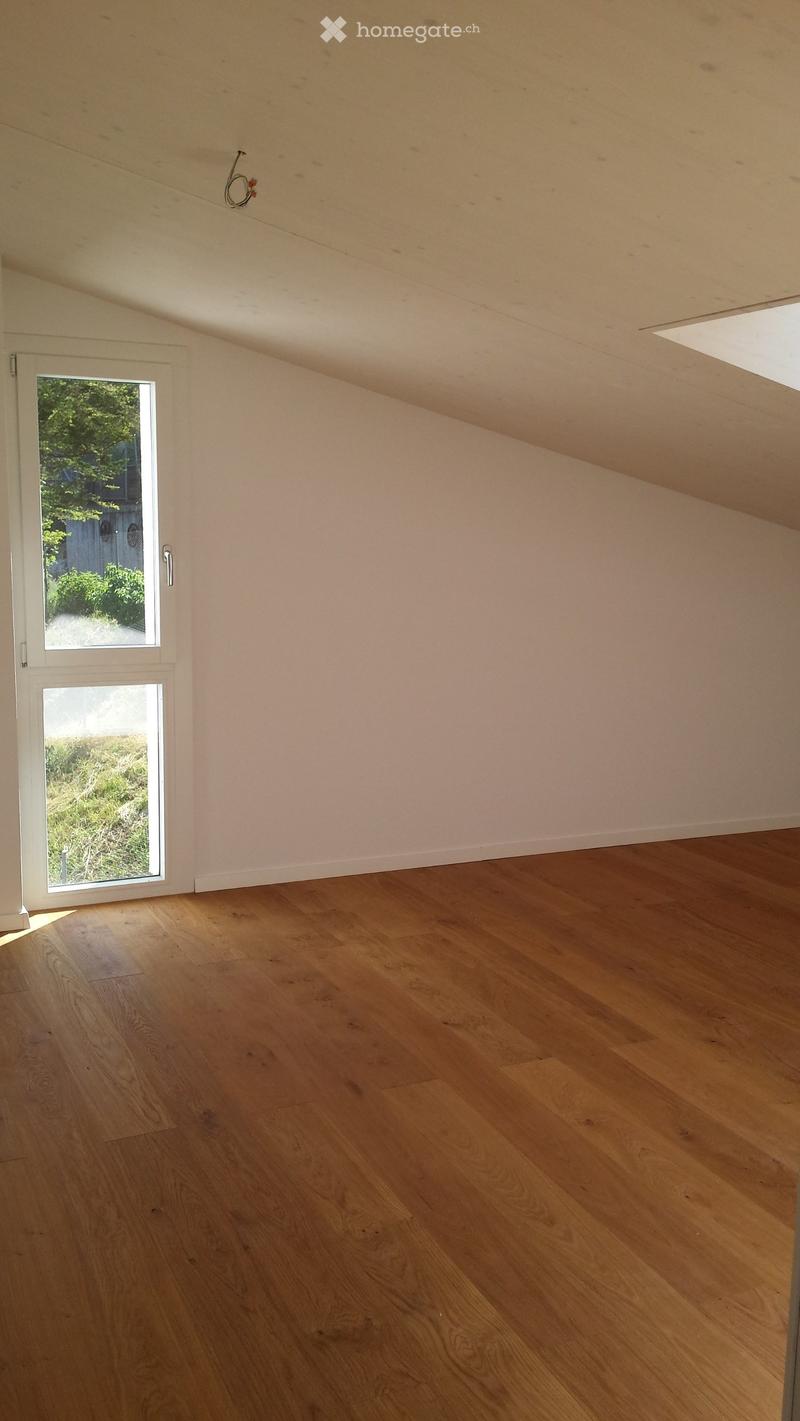 Empty room with wooden flooring, white walls, and a large window with a view of greenery outside.