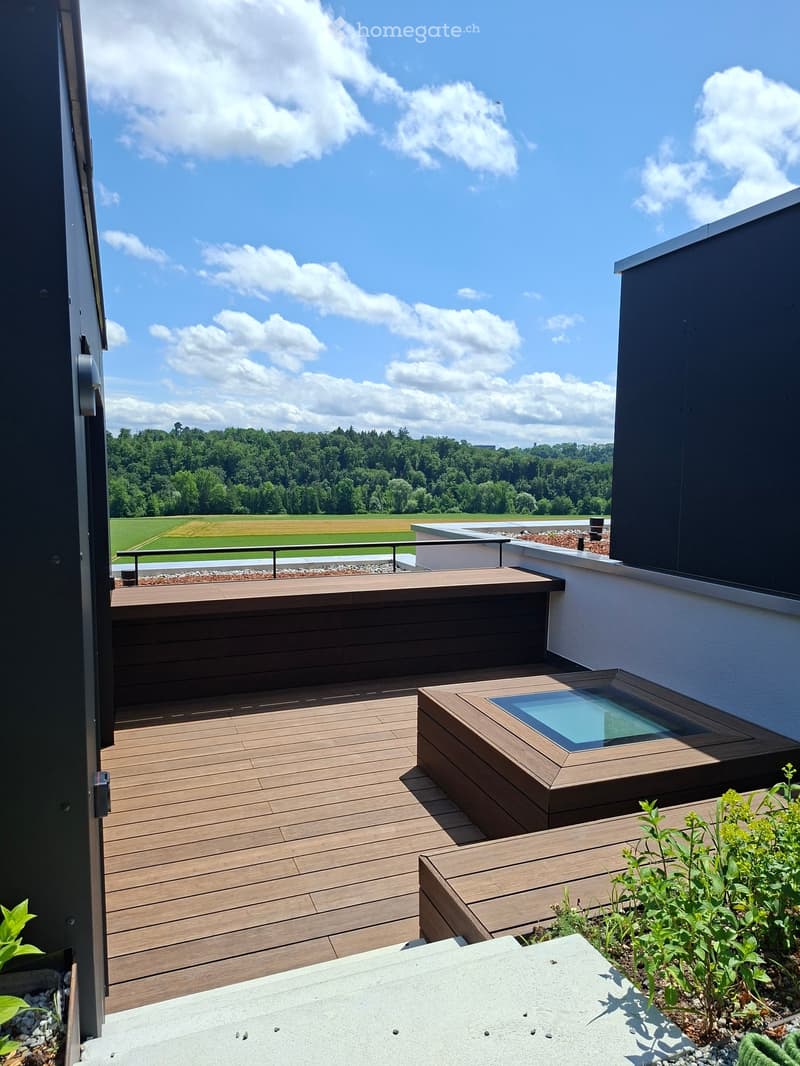 Wooden deck terrace with a glass-enclosed seating area, overlooking a green landscape with trees and fields.