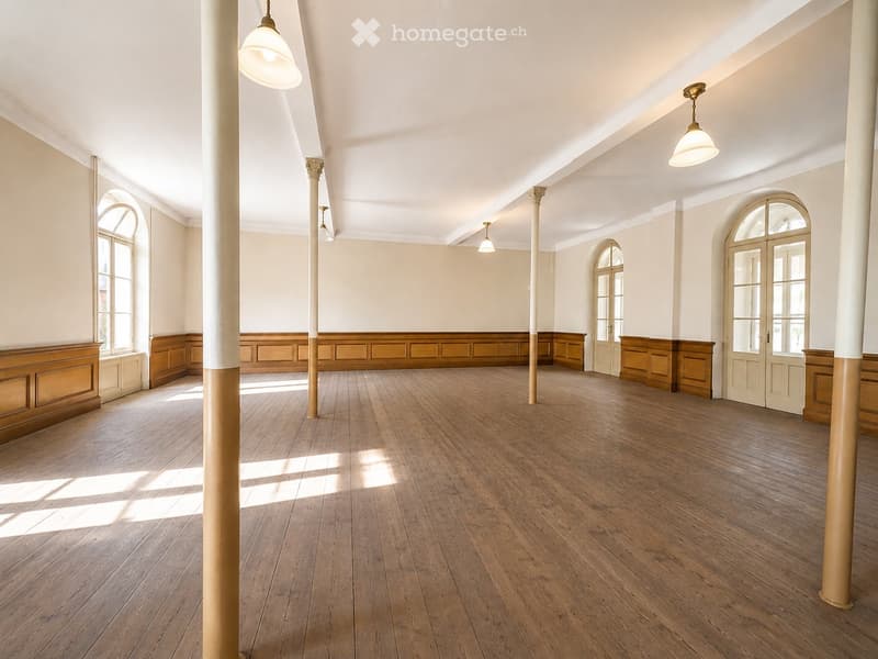Empty room with wooden flooring, white walls, large arched windows, and central columns. No furniture or equipment present.