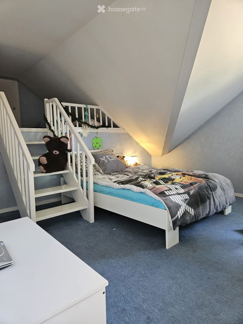 Cozy attic bedroom with a white bed, staircase leading to a loft area, and a small table. The room features blue carpeting and a slanted ceiling.