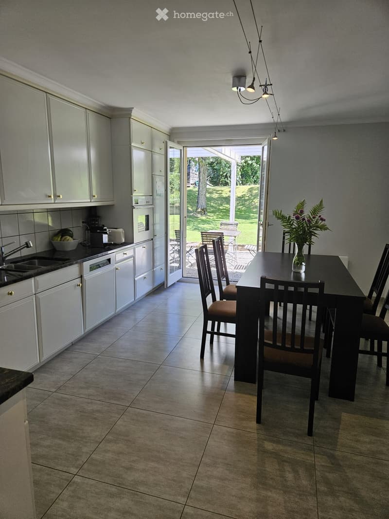 Open-plan kitchen and dining area with white cabinets, a dark dining table with chairs, and a vase of flowers on the table. The kitchen features a sink, oven, and dishwasher, with a view to a garden through large glass doors.