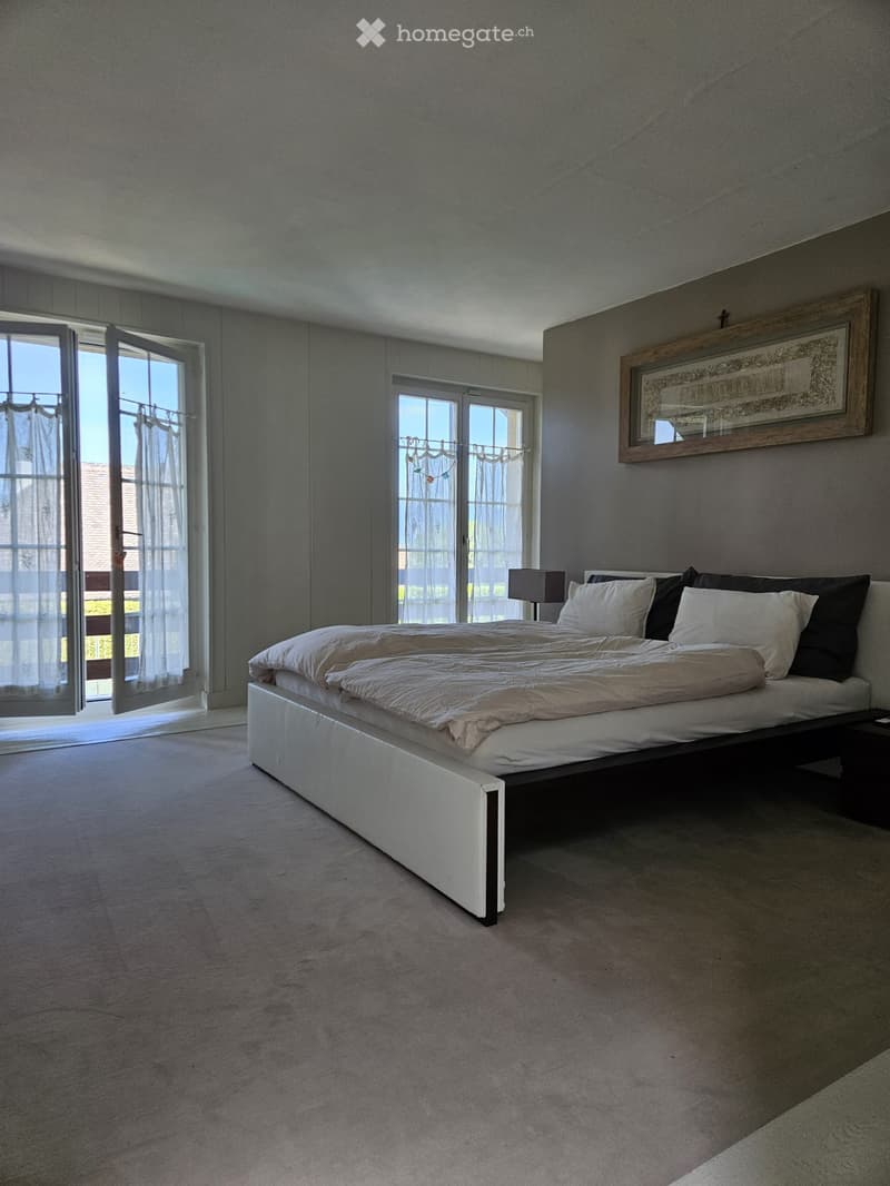 Modern bedroom with a large bed, nightstand, lamp, and picture frame on the wall. Large windows with sheer curtains allow natural light to fill the space.