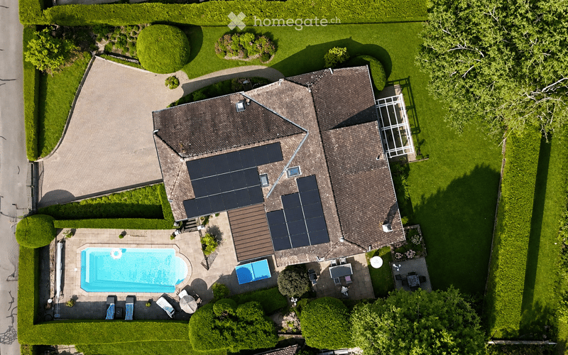 Aerial view of a two-story house with a tiled roof, solar panels, and a well-maintained garden. The property includes a swimming pool, patio furniture, and a driveway.