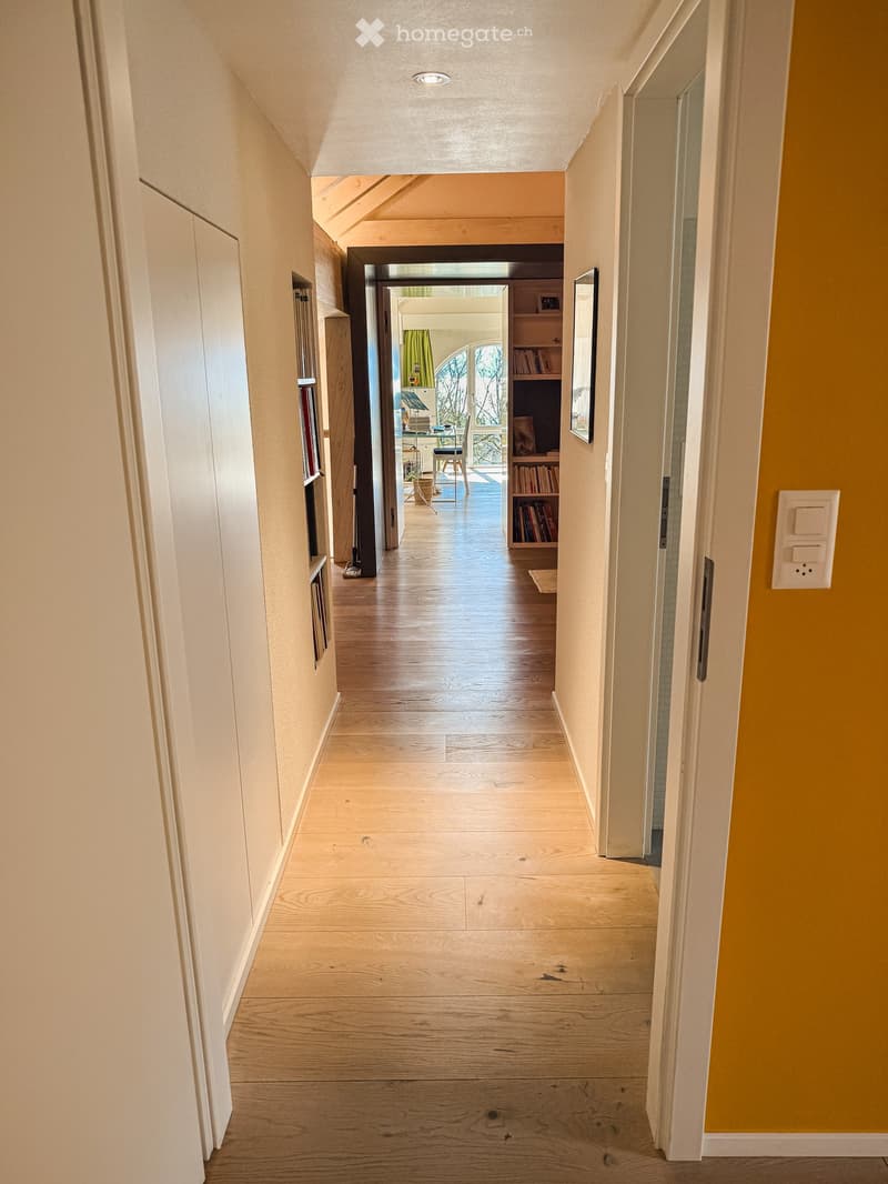 Bright hallway with wooden flooring, white walls, and recessed lighting. Visible rooms include a study with bookshelves and a desk, and a living area with a window and green curtains.
