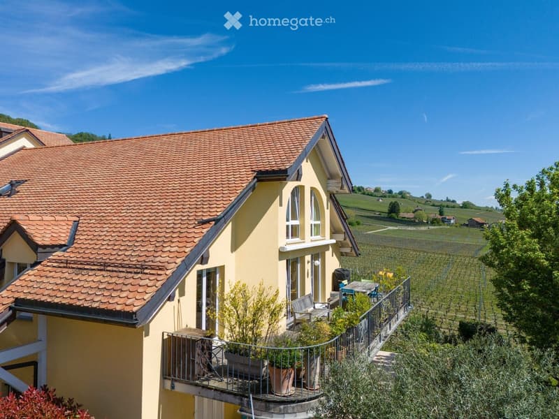 Two-story yellow house with red-tiled roof, large windows, and a balcony with outdoor furniture and potted plants.