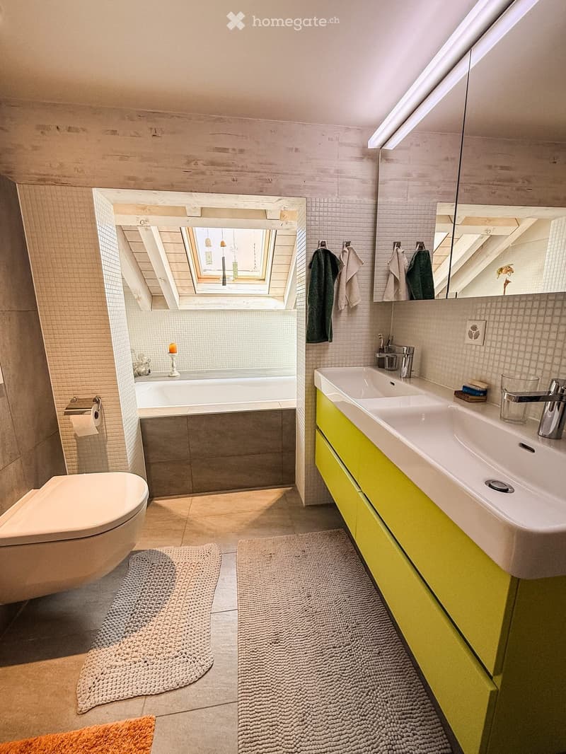 Modern bathroom with a bathtub, toilet, double sink, and large mirror. Features include a skylight window, bright yellow cabinets, and two round bath mats.