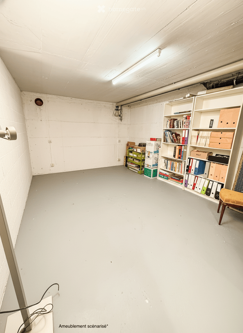 A spacious, minimally furnished room with white walls and a grey concrete floor. Features include a large bookshelf filled with books and storage boxes, a small wooden chair, and exposed pipes and electrical fixtures on the walls.