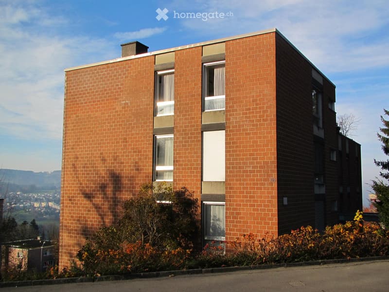 Modern two-story brick building with large windows and a flat roof, surrounded by greenery and shrubs.