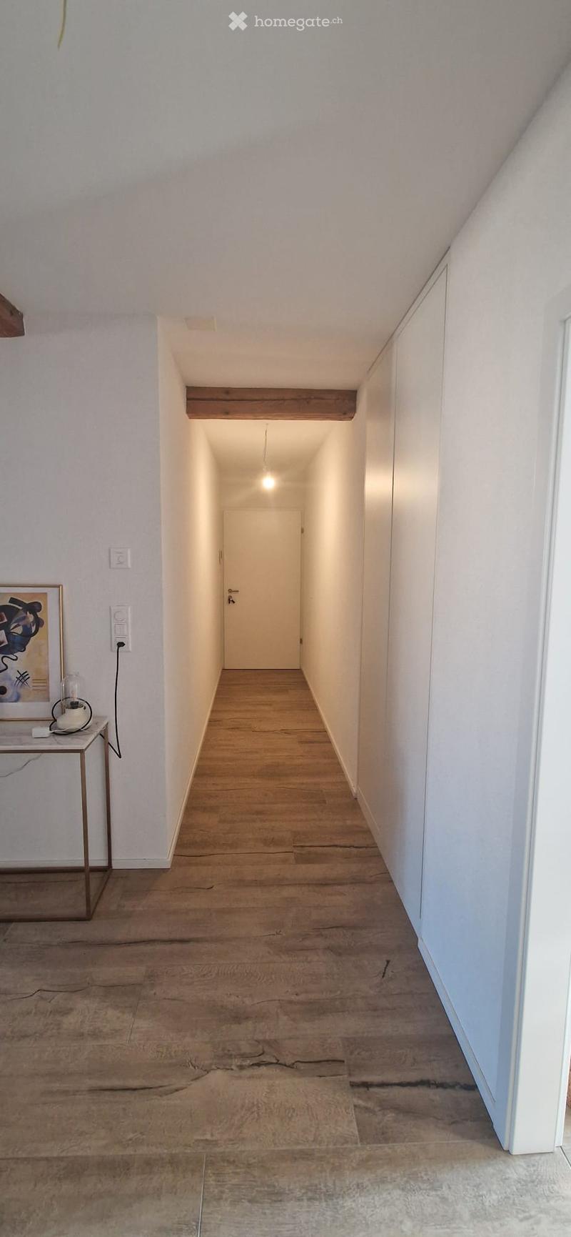 A narrow hallway with wooden flooring, white walls, and a single door at the end. A small table with a lamp and a framed picture is visible on the left side.