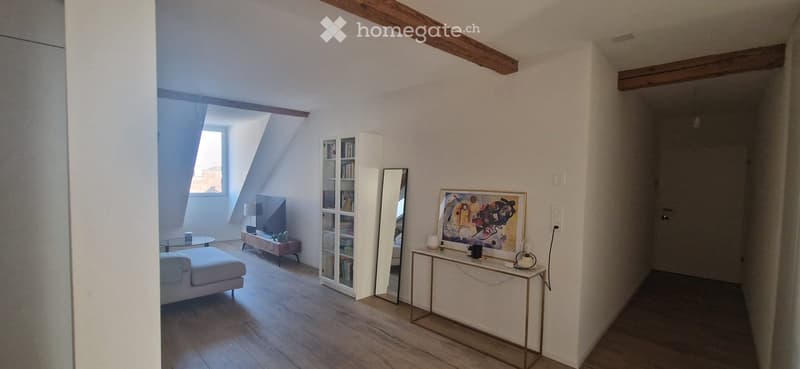 Bright living room with wooden floors, white walls, and exposed wooden beams. Features a white sofa, glass coffee table, bookshelf, mirror, and a small table with decorative items.