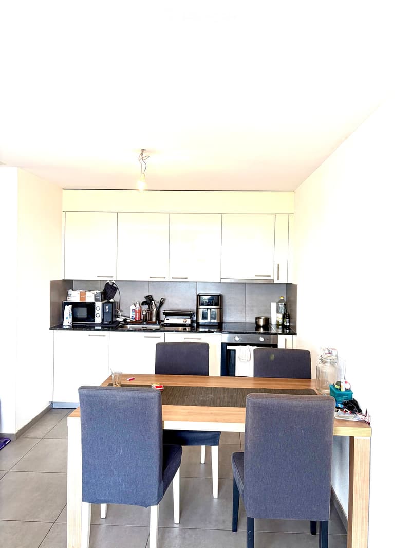 Modern kitchen with white cabinets, a wooden dining table with four chairs, and various appliances including a microwave, oven, and fridge.