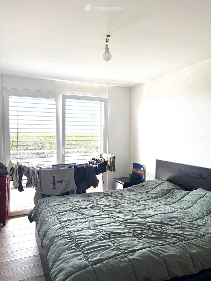 Bright bedroom with wooden floor, large windows with blinds, bed with green duvet, nightstand with picture frame, clothesline with hanging clothes, and a red suitcase near the window.