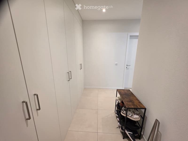 A narrow hallway with white walls, tiled floor, built-in wardrobes, and a small wooden table with a shoe rack holding multiple pairs of shoes.