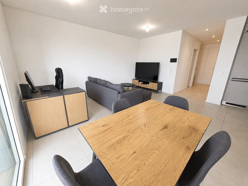 Modern living room with a grey sofa, wooden coffee table, chairs, TV, and a desk with a computer setup.