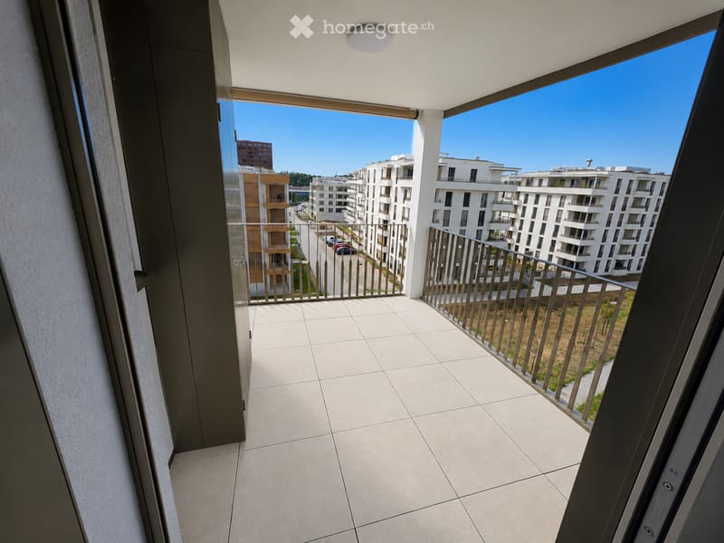 Modern balcony with tiled floor, metal railing, and expansive view of a residential area with contemporary buildings and clear blue sky.