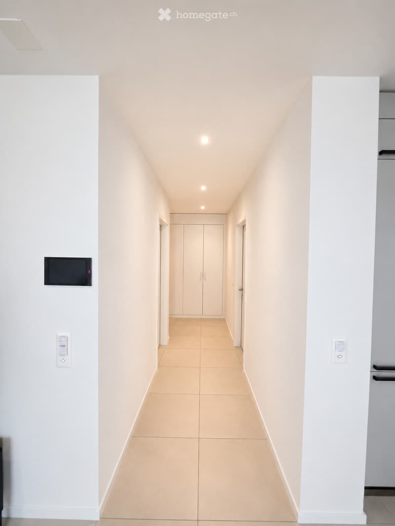 A clean, modern hallway with light-colored walls and tiled flooring, featuring recessed ceiling lights and a closed door at the end.