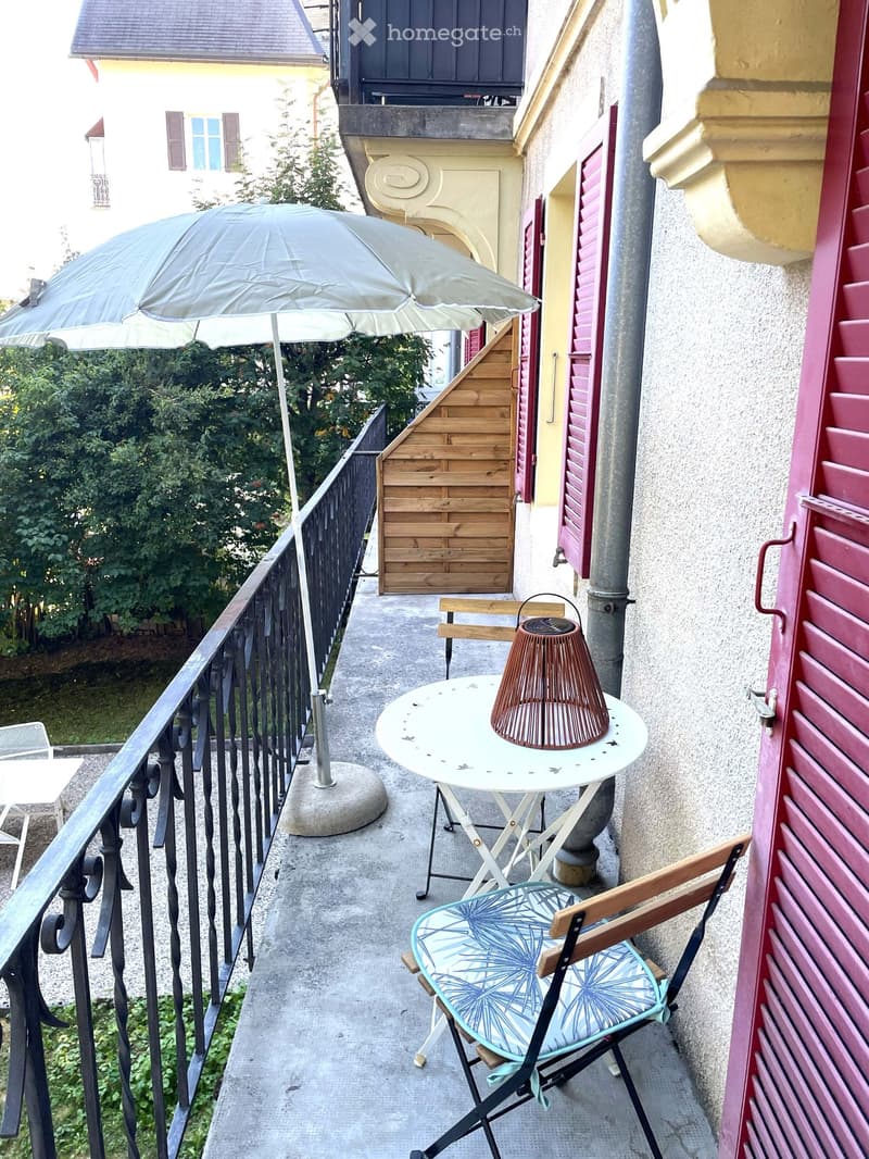 Balcony with a small round table, a wicker lamp, a folding chair with a blue patterned cushion, and an umbrella.
