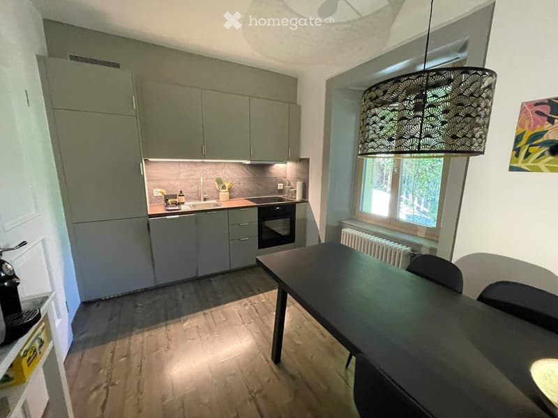 Modern kitchen with grey cabinets, a sink, oven, and a dining area with a table and chairs. A large window provides natural light, and a decorative lamp hangs from the ceiling.