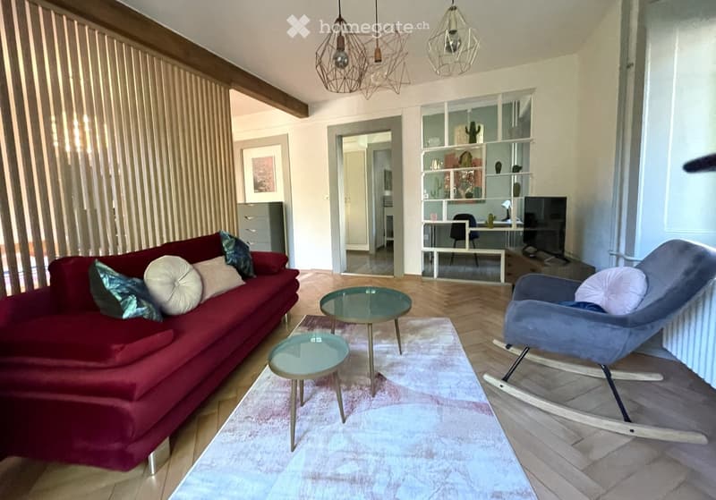 Modern living room with a red sofa, glass coffee tables, a blue armchair, and a shelf with decorative items.