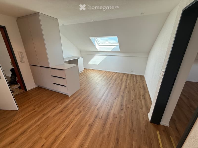 An empty attic room with wooden flooring, white walls, and a skylight window. The room features built-in white cabinets and drawers along one wall.
