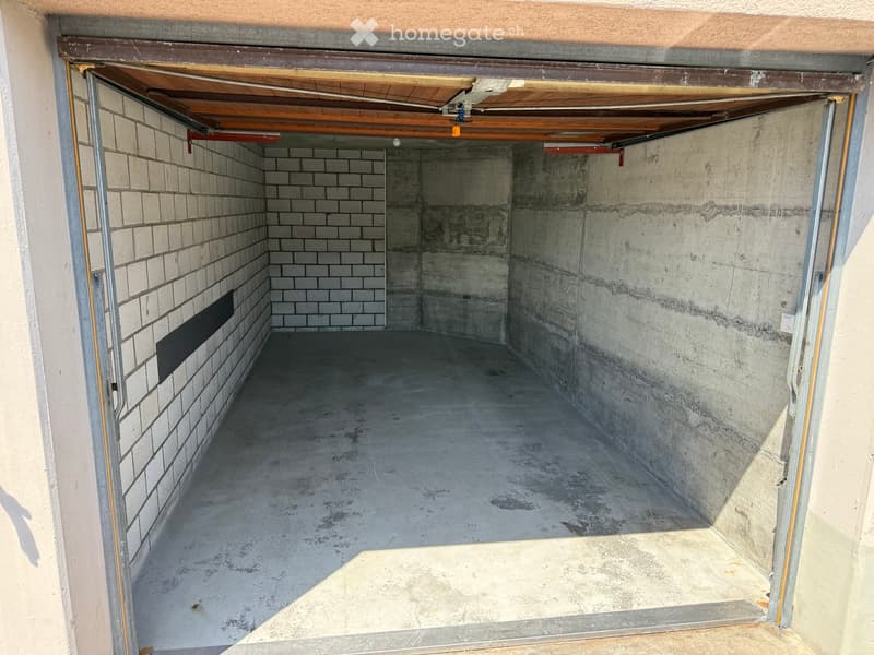 Empty garage with concrete floor, brick wall, and wooden ceiling beams. No furniture or equipment present.